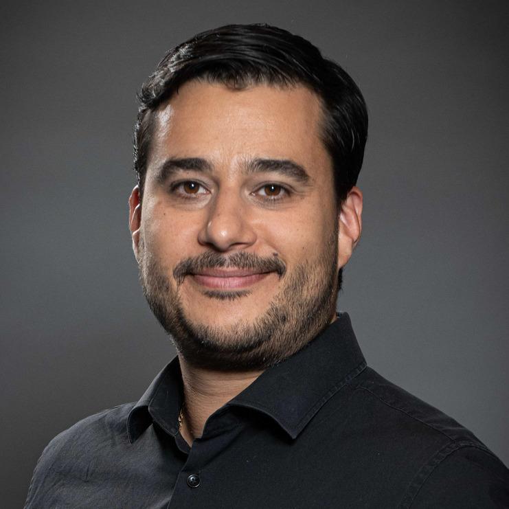 Porträt eines Mannes in schwarzem Hemd vor einem neutralen, dunklen Hintergrund, mit leicht lächelndem Gesichtsausdruck.