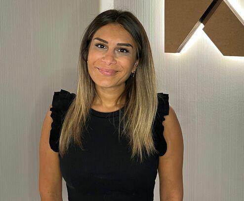 Frau mit langen, braunen Haaren steht vor einer Wand mit beleuchtetem, braunem Buchstaben "K".