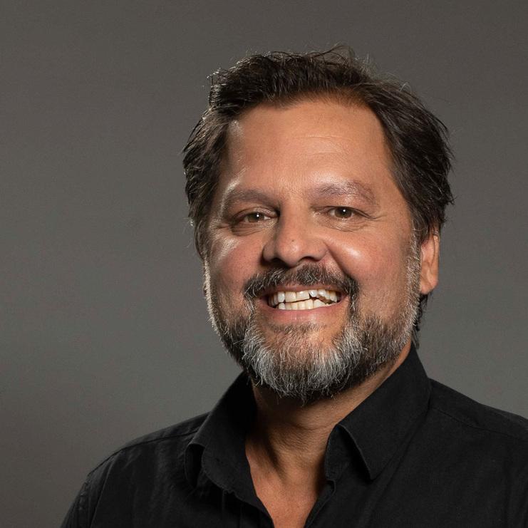 Mann mit Bart und kurzem, dunklem Haar, lächelt in schwarzem Hemd vor einfarbigem Hintergrund. Händen in den Taschen.