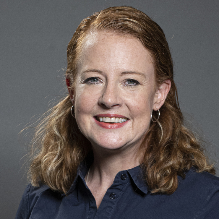 Portr&auml;t einer l&auml;chelnden Frau mit langen, lockigen Haaren, die ein dunkles Hemdkleid tr&auml;gt, vor grauem Hintergrund.