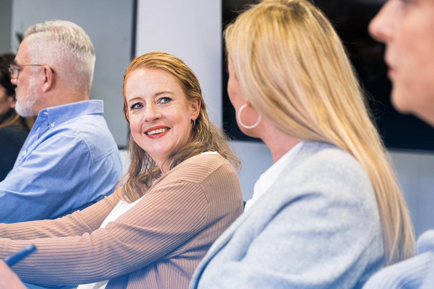 L&auml;chelnde Frau in einem Meeting, schaut zu einer anderen Frau, mehrere Teilnehmer sind im Hintergrund sichtbar.