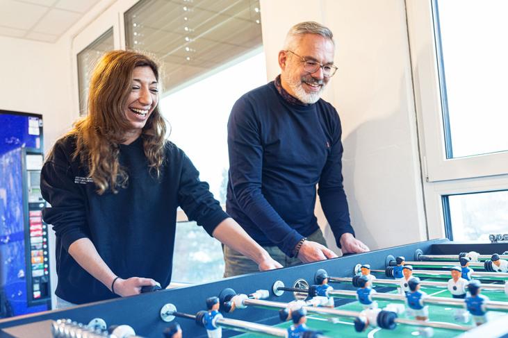 Zwei Personen spielen begeistert am Tischfußballkasten in einem hellen Raum mit Fenstern.