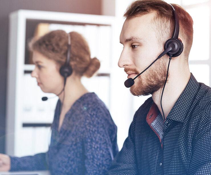 Zwei Personen mit Headsets sitzen an einem Tisch, konzentriert auf ihre Arbeit in einem modernen Büro.