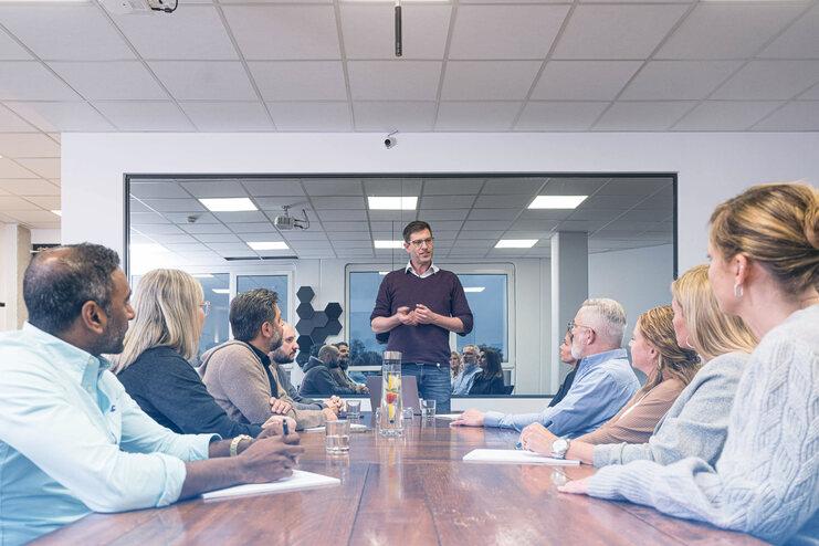 Mann steht vor einer Gruppe von sechs Personen in einem Konferenzraum, während er eine Präsentation hält.