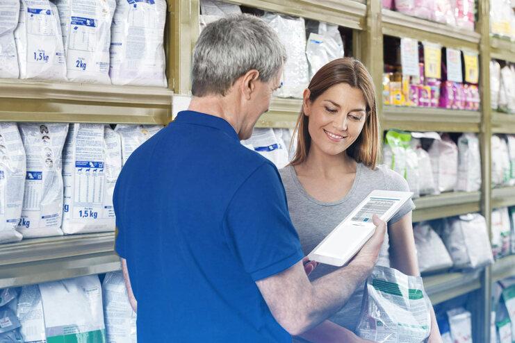 Ein Mann zeigt einer Frau auf einem Tablet Informationen zu Produkten im Hintergrund einer Tierfutterabteilung.
