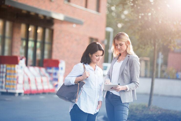 Zwei Frauen betrachten gemeinsam ein Tablet vor einem Gebäude, umgeben von Einkaufskörben im Hintergrund.