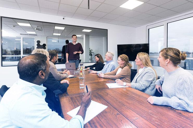 Mitarbeiter sitzen an einem Tisch, w&auml;hrend ein Kollege mit Laptop eine Pr&auml;sentation im Meetingraum h&auml;lt.