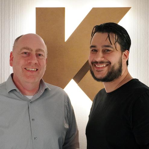 Zwei Männer lächeln in einem Innenraum vor einer beleuchteten, stilisierten „K“-Wand.