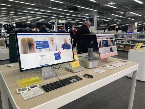 Zwei Computer an einem Verkaufstisch in einem Elektromarkt, einer mit Präsentation, der andere im Hintergrund sichtbar.
