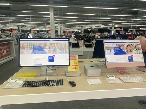 Zwei Computerbildschirme mit Produktinformationen in einem Elektronikgeschäft, im Vordergrund ein Mac mini und Zubehör.