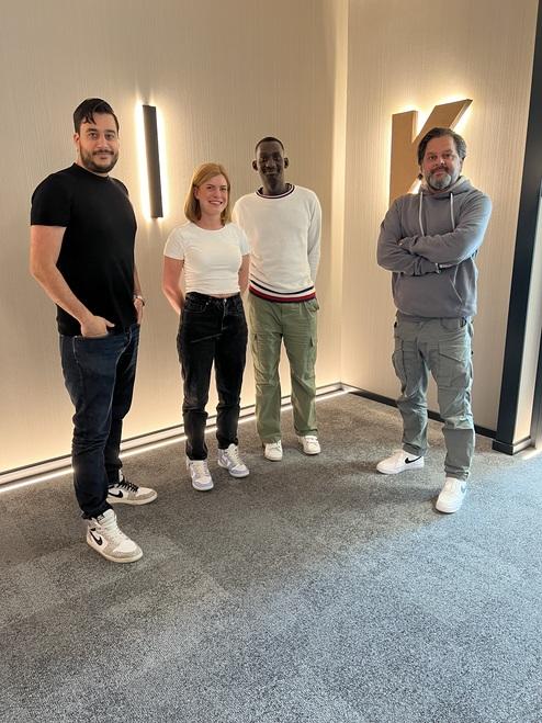 Vier Personen stehen lächelnd vor einer beleuchteten Wand mit dem Buchstaben "K" in einem modernen Raum.