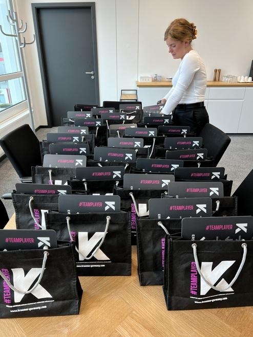 Frau steht neben einem Tisch, umgeben von mehreren schwarz-beigen Taschen mit dem Hashtag „#TEAMPLAYER“.