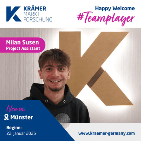 Junger Mann mit dunklem Hoodie lächelt vor einer beleuchteten, großen K-formigen Wanddekoration.