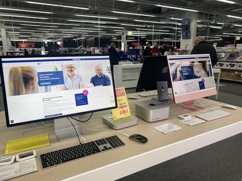 Zwei Computer mit Webseiten in einem Elektronikgeschäft, kombiniert mit Zubehör auf einem Holztisch.
