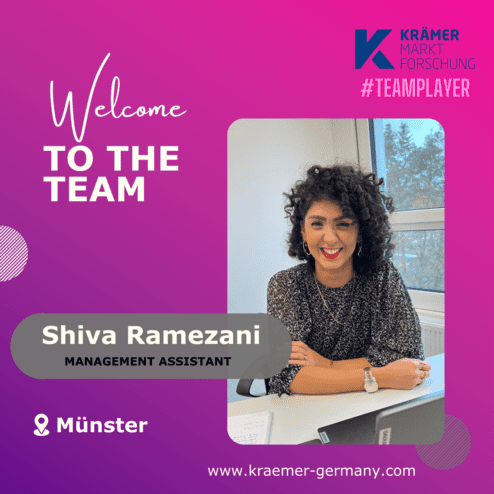 Frau mit lockigem Haar lächelt am Schreibtisch, umgeben von Dokumenten und einem Laptop, Hintergrund mit Fenster.