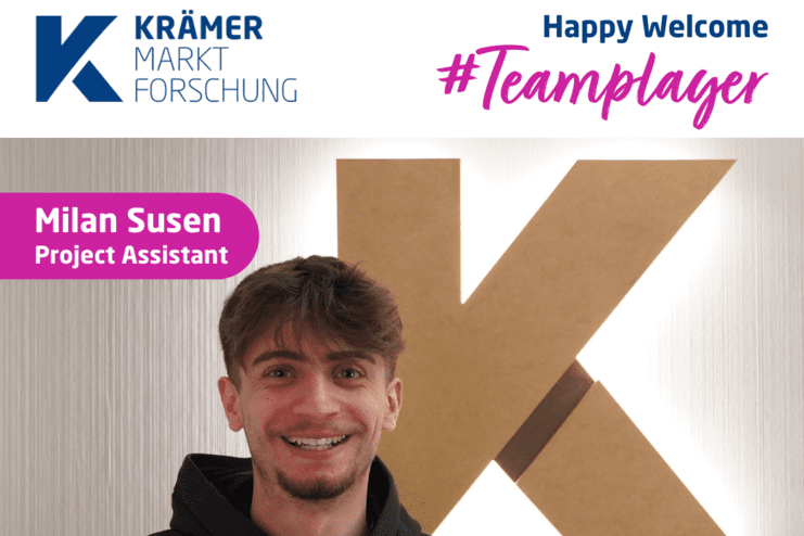 Junger Mann mit dunklem Hoodie lächelt vor einer beleuchteten, großen K-formigen Wanddekoration.