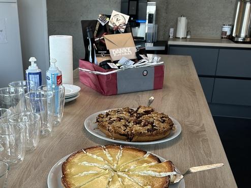 Zwei Kuchen auf einem Tisch: ein geschnittenes Käsekuchenstück und ein Schokoladenkuchen, umgeben von Gläsern und Geschirr.