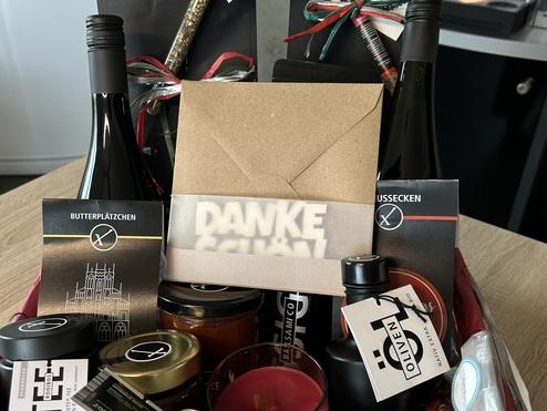 Geschenkkorb mit Wein, Keksen, einem Candle Jar, Marmelade und weiteren Delikatessen auf einem Holztisch.