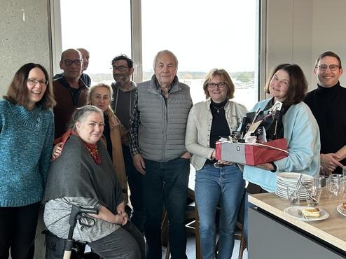 Gruppe von neun Personen in einem Raum, einige lächeln, andere halten Geschenke, eine Person sitzt im Rollator.