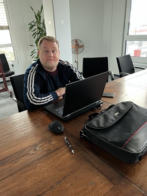 Mann mit kurzem Bart sitzt an einem Tisch mit Laptop, Maus und Aktenmappe in einem Büro. Im Hintergrund ein Pflanze.