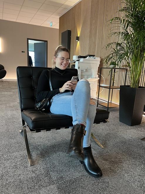 Junge Frau sitzt lächelnd auf einem schwarzen Sessel und schaut auf ihr Smartphone in modernem Büroambiente.