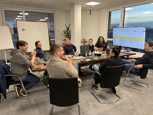 Teammeeting in einem modernen Büro mit acht Personen, Laptop und Projektor auf einem Tisch. Hintergrund mit Fenstern.