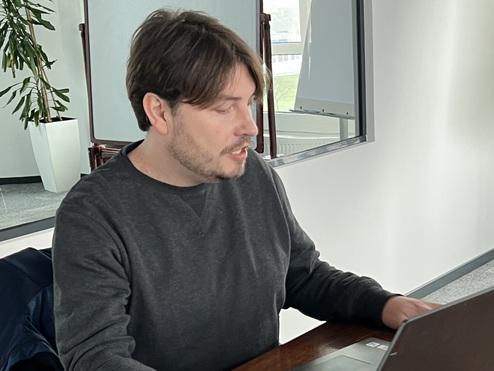 Mann sitzt an einem Tisch mit einem Laptop und einer Tasse Kaffee, umgeben von hellen Büroräumen.