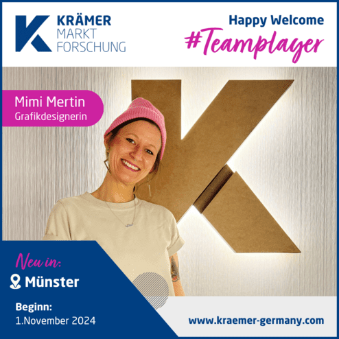 Frau mit pinker Mütze und heller Kleidung lächelt vor einer beleuchteten Wand mit dem Buchstaben K von Krämer Markt Forschung.