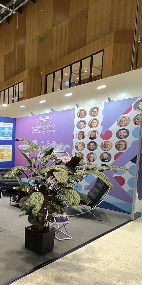 Messestand mit Loungemöbeln, Pflanzen und bunten Displays in einem Veranstaltungssaal, Menschen im Hintergrund.