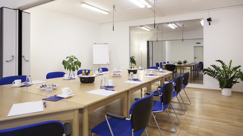 Konferenzraum mit langem Tisch, blauen Stühlen, Flipchart, Pflanzen und Getränken in Gläsern auf dem Tisch.