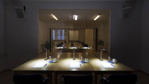 Leerer Besprechungsraum mit einem langen Tisch, Stühlen und rechteckiger Leinwand, beleuchtet durch Schreibtischlampen.