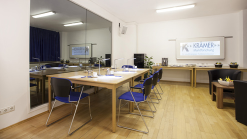 Moderne Besprechungsraum mit Holz-Tisch, blauen Stühlen, Präsentationsbildschirm und Snackbereich.