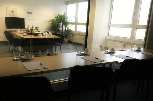 Konferenzraum mit einem großen Tisch, Stühlen, Getränken und Notizblöcken, im Hintergrund Fenster und Pflanzen.